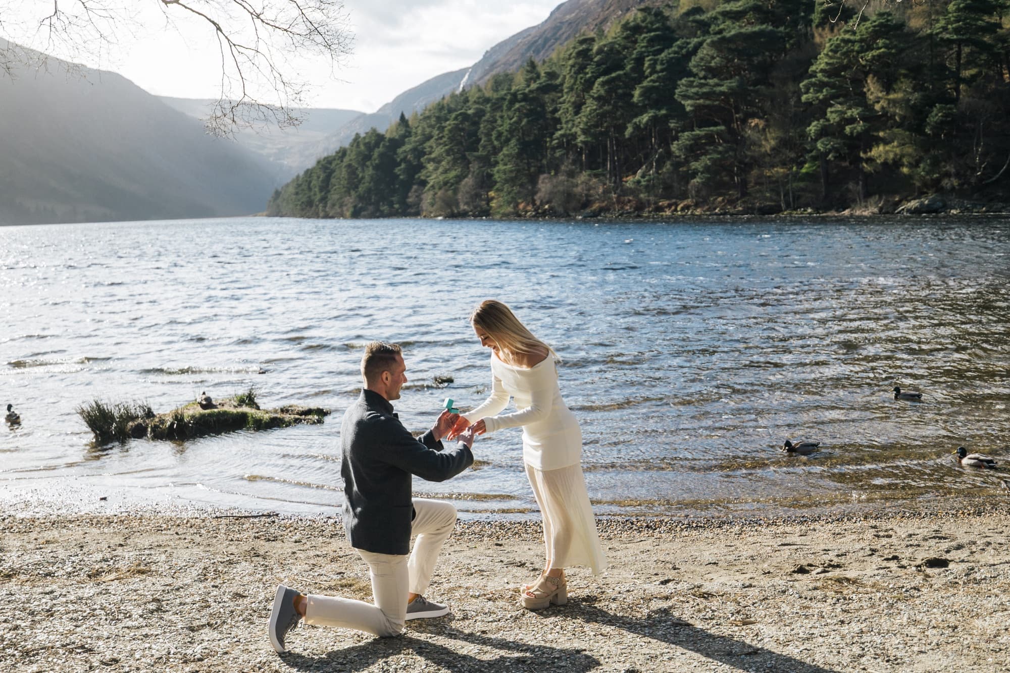 Matt on one knee on the shore of the lake at Glendalough, holding a ring out to Anna, with the mountains behind.